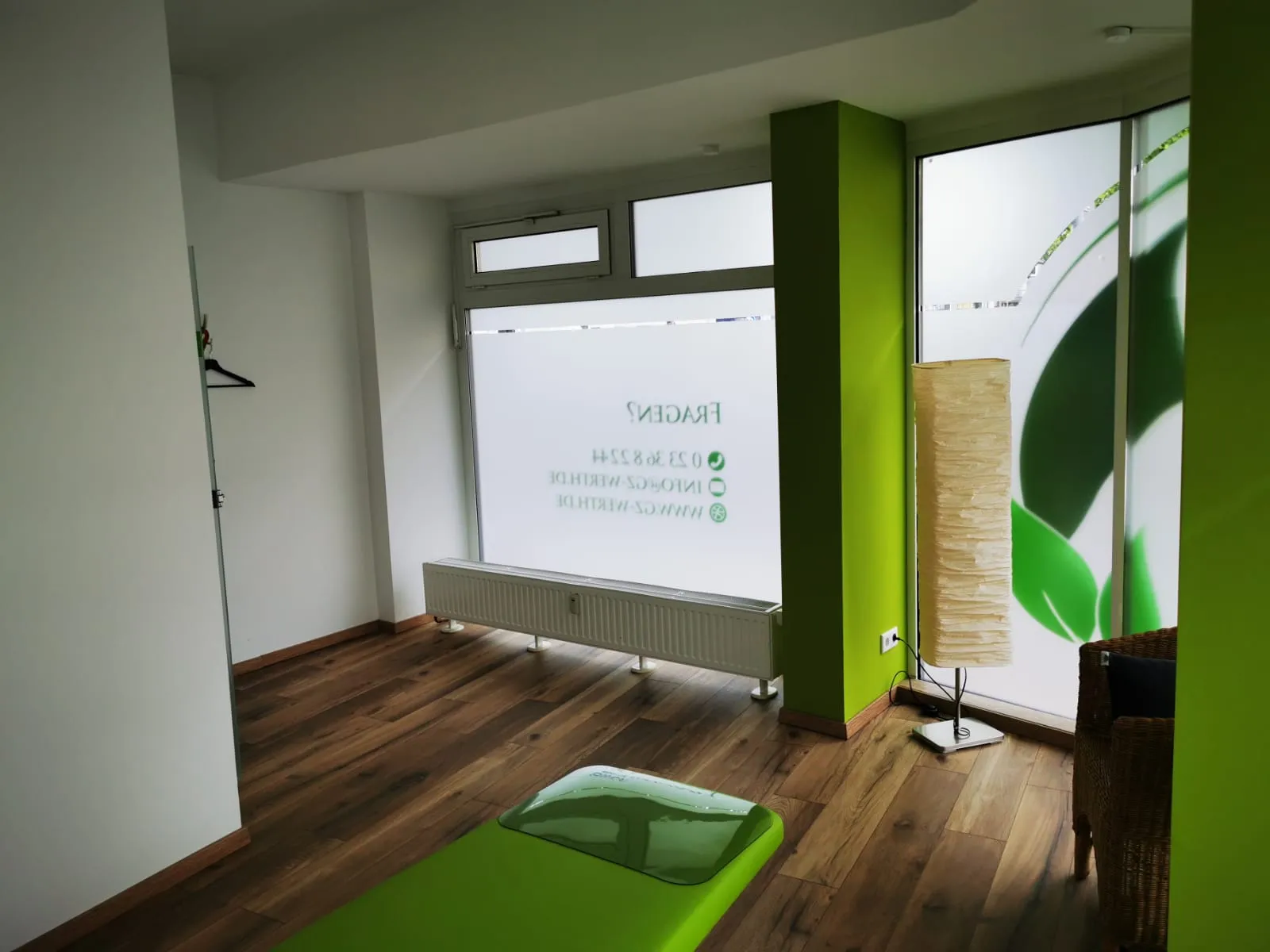 Gesundheitszentrum Werth - Physiotherapie und medizinische Behandlungen - Ein heller, moderner Therapieraum mit einer grünen Massageliege auf Holzboden, einem großen Fenster mit mattierten Geschäftsdetails, einer hohen Lampe und grünen Wandakzenten.